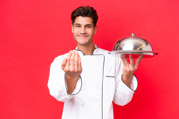 Νεαρός καυκάσιος σεφ κρατά το δίσκο απομονωμένο σε κόκκινο φόντο προσκαλώντας τον να έρθει με το χέρι. Χαίρομαι που ήρθες. - Φωτογραφία, εικόνα