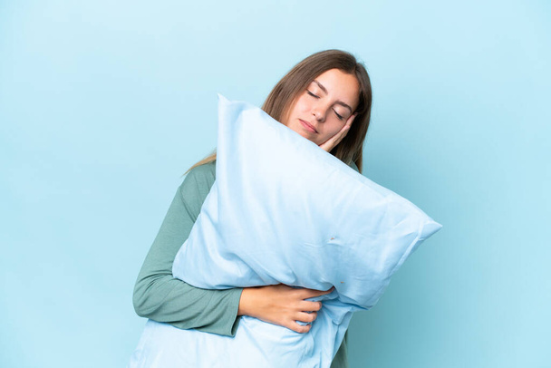 Giovane bella donna isolata su sfondo blu in pigiama e tenendo un cuscino mentre dorme - Foto, immagini