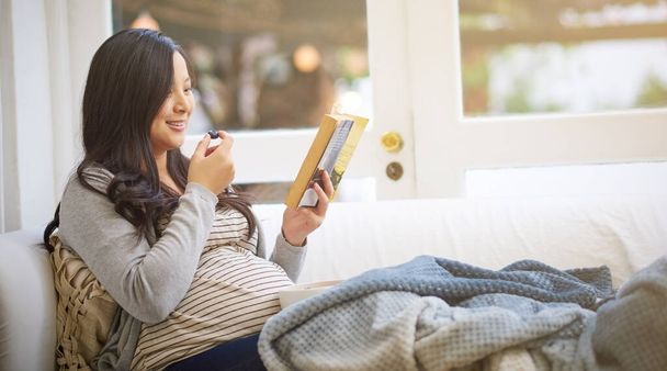 Το μωρό της τρελαίνεται όταν τρώει οργανική τροφή. μια ελκυστική νεαρή έγκυος γυναίκα τρώει φρούτα και διαβάζει ένα βιβλίο ενώ χαλαρώνει στον καναπέ στο σπίτι - Φωτογραφία, εικόνα