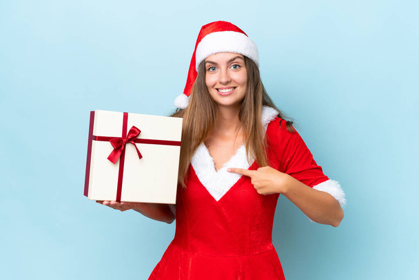 Anne Noel gibi giyinmiş genç beyaz bir kadın elinde hediyeyle mavi arka planda, sürpriz bir yüz ifadesiyle izole edilmiş. - Fotoğraf, Görsel