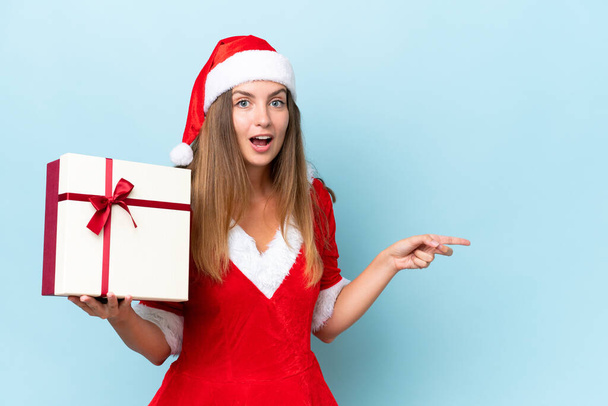 Anne Noel gibi giyinmiş, mavi arka planda izole edilmiş bir hediye taşıyan beyaz bir kadın şaşırmış ve parmağıyla kenarı işaret ediyor. - Fotoğraf, Görsel