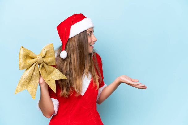 Young caucasian woman dressed as mama noel holding Christmas bow isolated on blue background with surprise facial expression - Fotó, kép
