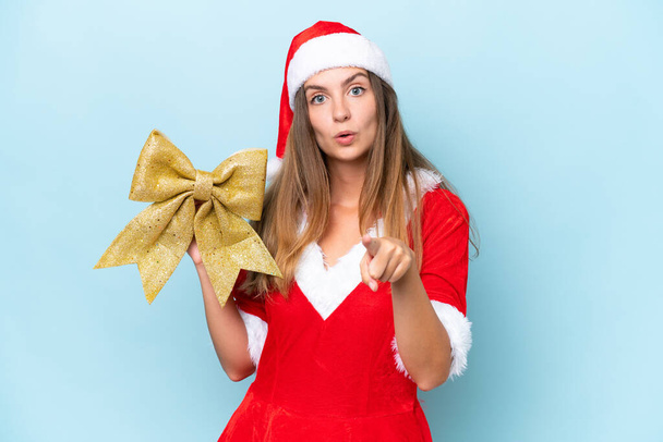 Anne Noel gibi giyinmiş, elinde Noel yayı tutan beyaz bir kadın mavi arka planda izole edilmiş, şaşırmış ve ön tarafı işaret ediyor. - Fotoğraf, Görsel