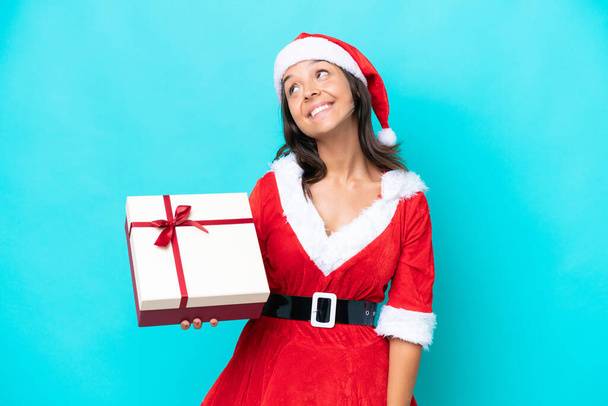 Anne Noel gibi giyinmiş genç İspanyol kadın elinde mavi arka planda izole edilmiş bir hediyeyle yukarı bakarken bir fikir düşünüyor. - Fotoğraf, Görsel