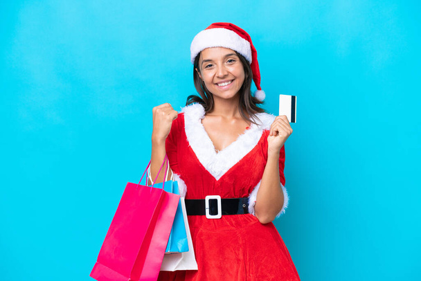 Anne Noel gibi giyinmiş genç İspanyol kadın mavi arka planda izole edilmiş. Elinde alışveriş çantaları ve bir kredi kartı var. - Fotoğraf, Görsel