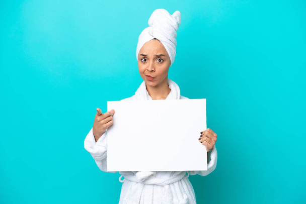 Young woman in a bathrobe with towel isolated on blue background holding an empty placard and pointing to the front - Zdjęcie, obraz