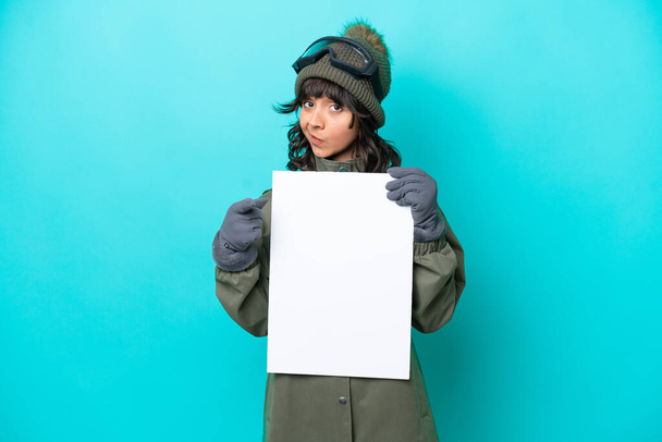 Лыжная латиноамериканка в очках для сноуборда на синем фоне держит пустой плакат и указывает на фронт - Фото, изображение