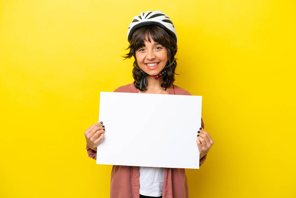 Young cyclist latin woman isolated on yellow background holding an empty placard with happy expression - Foto, immagini