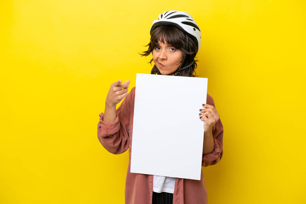 Молода жінка-велосипедистка латинка ізольована на жовтому тлі, що тримає порожній плакат і вказує на передній план
 - Фото, зображення