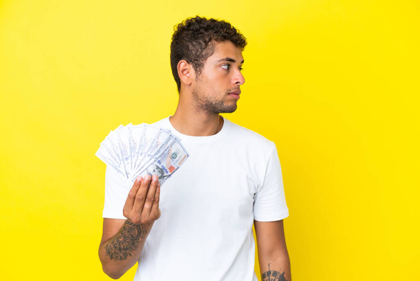 Fiatal brazil férfi vesz egy csomó pénzt elszigetelt sárga háttér néz oldalra - Fotó, kép