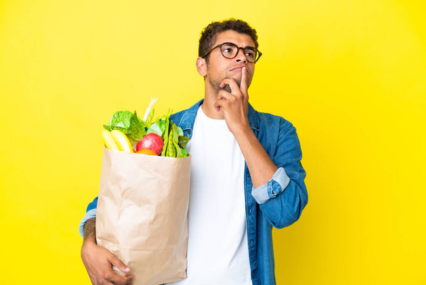 Young Brazilian man holding a grocery shopping bag isolated on yellow background having doubts while looking up - Foto, Bild