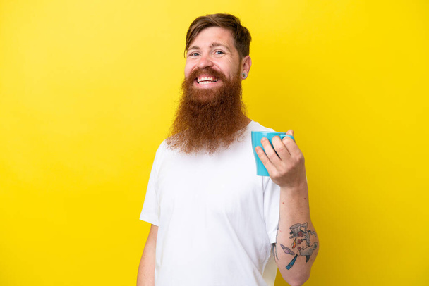 Vörös hajú férfi szakállal, kezében egy bögre elszigetelt sárga háttérrel, sokat mosolyog. - Fotó, kép