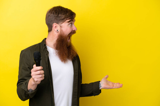 Pelirrojo con barba recogiendo un micrófono aislado sobre fondo amarillo con expresión facial sorpresa - Foto, Imagen
