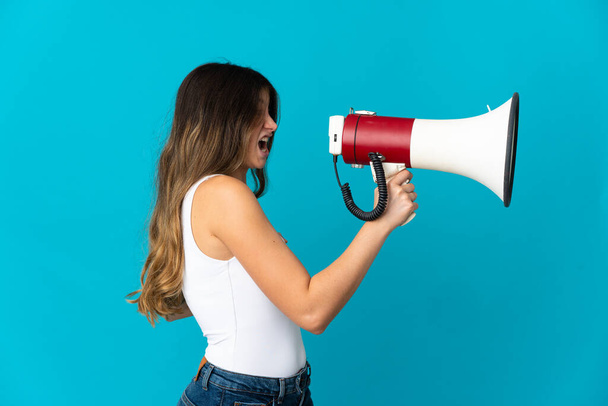 Young caucasian woman isolated on blue background shouting through a megaphone to announce something in lateral position - Foto, imagen