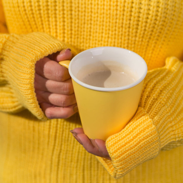 Női kéz tart egy sárga csésze egy cappuccino ital, a háttérben egy sárga pulóver, koncepció, szelektív fókusz - Fotó, kép