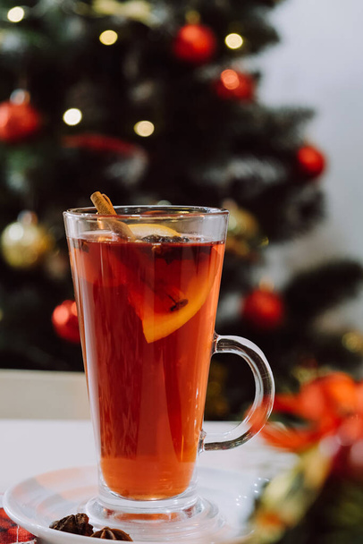 Tarçınlı ve portakal meyveli sıcak şarap. Arka planda süslenmiş Noel ağacının üzerindeki beyaz masada tatil için sunulan ılık kış içeceğinin dikey görüntüleri. Mutlu yıllar konsepti. Seçici odak - Fotoğraf, Görsel