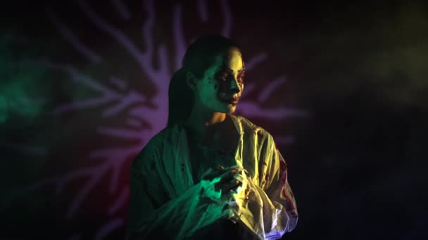 Jonge vrouw portretteert bloeddorstige zombie met horror wonden op haar gezicht en bloederige kleren tegen donkere achtergrond met verlicht. Enge afbeelding voor Halloween. - Video