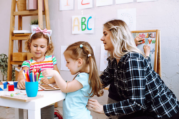 Χαρωπά κοριτσάκια διπλώνουν χρωματιστούς κύβους στο γραφείο στο playroom του νηπιαγωγείου. Νεαρός εκπαιδευτικός κάθεται με τα παιδιά στο τραπέζι στο πάτωμα και να βοηθήσει να παίξει μάθησης παιχνίδια πλευρά άποψη. Μάθημα βρεφονηπιακού σταθμού. - Φωτογραφία, εικόνα