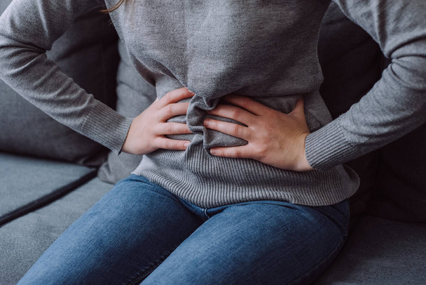 Close up of sick young female suffering stomach pain from food poisoning sitting on grey sofa at home. Unhealthy girl holding belly suffering from abdomen tummy pain. Woman gastritis health problem - 写真・画像