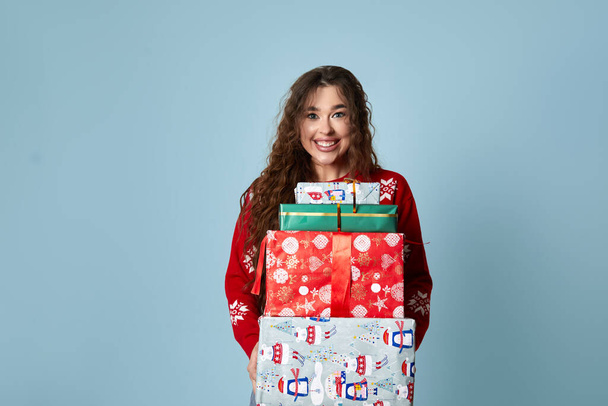 Happy woman red sweater holding boxes. Girl with gifts in hand smiling over blue background - Foto, afbeelding