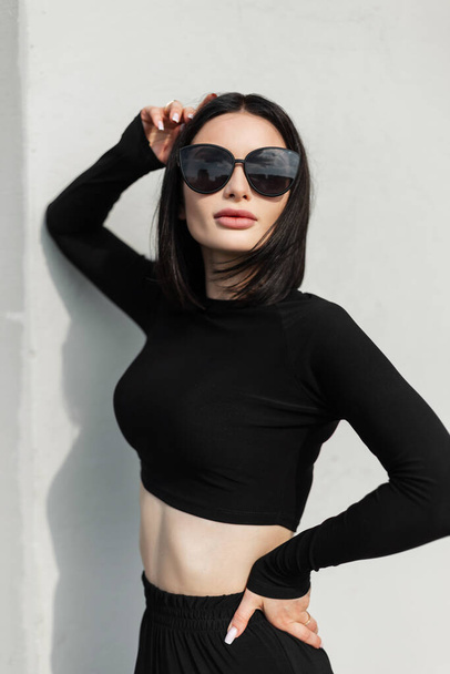Divatos városi stílusos nő hűvös napszemüveg fekete sportruházat a városban, közel a szürke fal. Női utcai ruhák néz ki - Fotó, kép