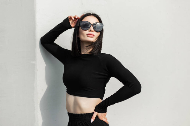 Moda cool girl w stylowych modnych okularach przeciwsłonecznych w modne czarne ubrania z długim rękawem top stoi w pobliżu szarej ściany w słoneczny dzień. Kobiecy strój sportowy miejski - Zdjęcie, obraz