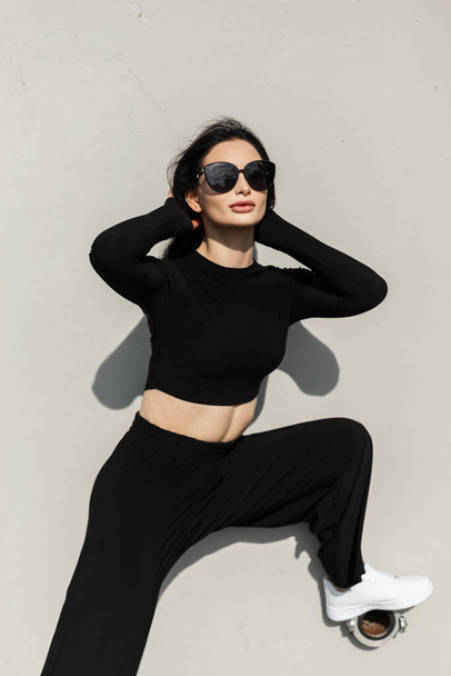 Модна красива жінка модель з модними сонцезахисними окулярами в модному чорному одязі з білими туфлями стоїть і позує біля сірої стіни на вулиці
 - Фото, зображення