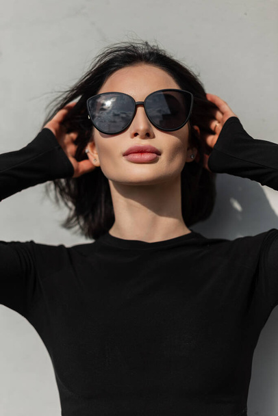 Retrato ensolarado urbano das mulheres de uma bela mulher fresca com lábios sexy na moda óculos pretos moldura na moda roupas pretas fica na luz do sol perto da parede cinza na rua - Foto, Imagem
