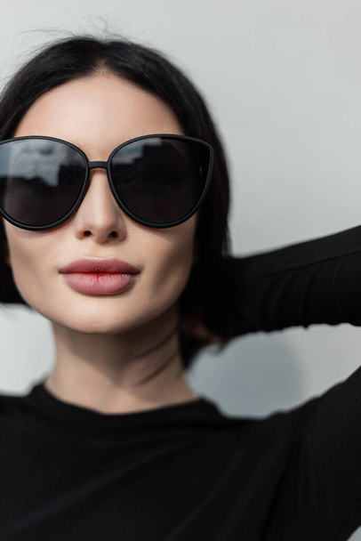 Cool female urban fresh portrait of a beautiful stylish girl with sexy lips wearing fashionable black sunglasses in a black T-shirt poses and fixes her hair near a gray wall in the city - Фото, изображение