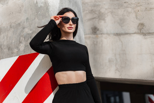 Stylish beautiful model girl in fashionable black sportswear puts on trendy sunglasses in the city near a concrete wall with red white lines - Photo, Image