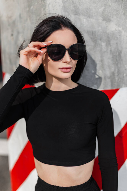 Portrait féminin urbain d'une belle fille élégante avec une coiffure bob dans des vêtements noirs à la mode met des lunettes de soleil à la mode et se tient près d'un mur en béton avec des lignes rouge-blanc - Photo, image