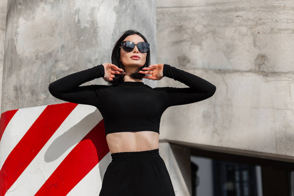 Modelo de menina elegante bonito elegante elegante com óculos de sol de moda legal em roupas pretas na moda na rua perto da parede de concreto com listras brancas e vermelhas - Foto, Imagem