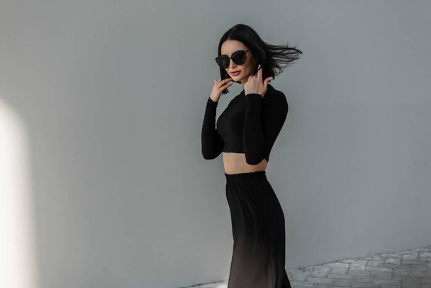 Retrato urbano femenino de la moda modelo de mujer hermosa con peinado en traje deportivo callejero negro con gafas de sol de moda negro camina en la ciudad cerca de una pared gris - Foto, Imagen