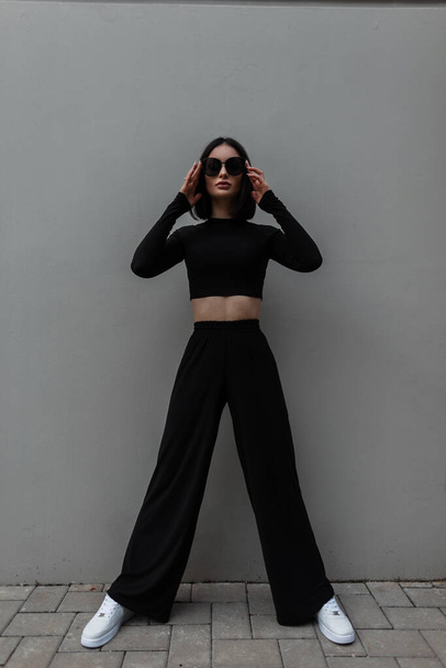 Cool stylish young woman with stylish black clothes with a long sleeve top and flared pants with white sneakers wears trendy sunglasses on a gray background - Foto, afbeelding