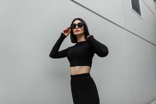 Cool fashion trendy beautiful woman in black fashionable clothes with top and pants wears stylish black sunglasses walks on the street near a modern gray building - Fotó, kép