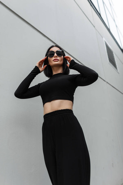 Schöne Frau in modischer schwarzer Kleidung trägt stylische schwarze Sonnenbrille und spaziert durch die Stadt in der Nähe eines modernen grauen Gebäudes - Foto, Bild