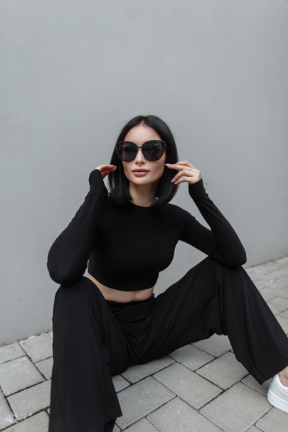 Модна міська красива жінка з чорними сонцезахисними окулярами в стильному спортивному вбранні сидить біля сірої будівлі на вулиці
 - Фото, зображення
