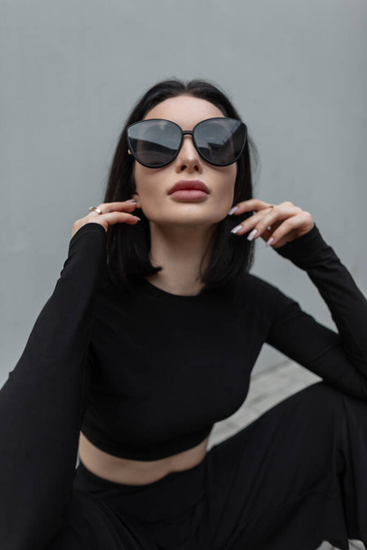 Stylowa piękna młoda kobieta z modą czarne okulary przeciwsłoneczne i modne czarne ubrania siedzi na ulicy w pobliżu szarej ściany - Zdjęcie, obraz