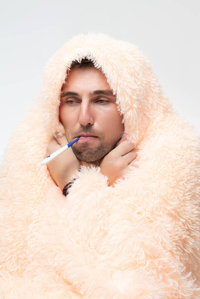 A sick man wrapped in a blanket holds a thermometer in his mouth, healthcare and treatment. - Photo, Image