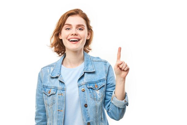 Portrait of positive redhead young woman in casual jeans smiling pointing with finger at empty copy space for text or product isolated on white background, advertising banner - Foto, Imagem