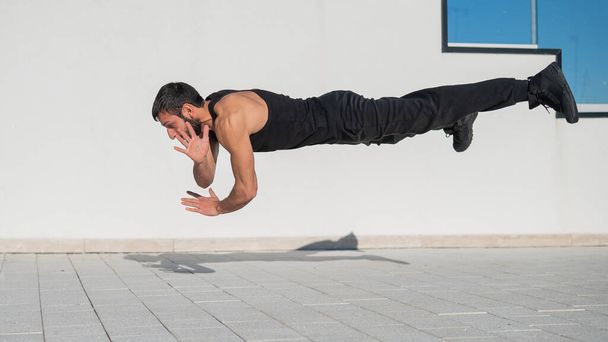 Un hombre en ropa deportiva negra salta mientras hace flexiones al aire libre - Foto, imagen