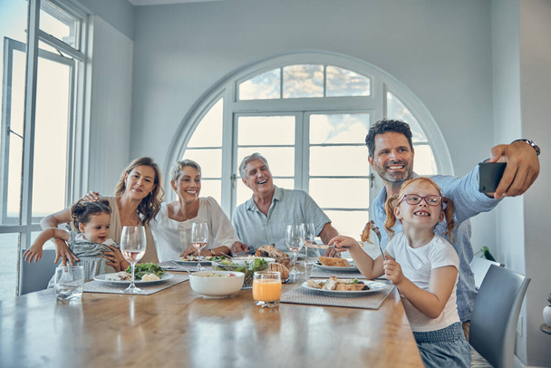 Бабушка с дедушкой, родители и дети делают селфи за ужином, обедают вместе по выходным. Большая семья, любовь и папа держат смартфон для фото наслаждаясь обедом, ужином и праздником. - Фото, изображение