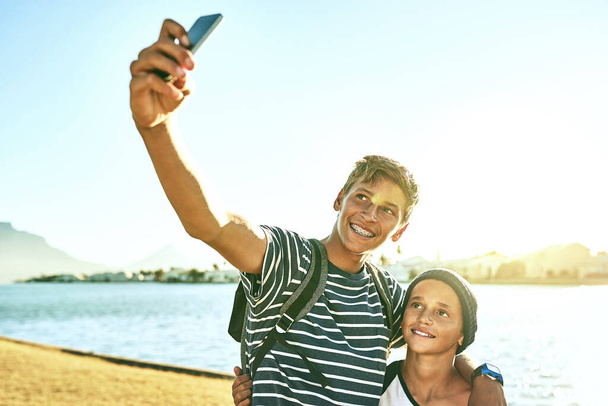 Tomarse una selfie bajo el sol. dos hermanos jóvenes tomando selfies afuera con una laguna en el fondo - Foto, imagen