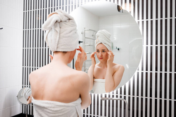 ホームバスルームで鏡の後ろに立つ抗しわクリームを適用する若い女性。美容と美容の手順。クレンジング後のスキンケア - 写真・画像