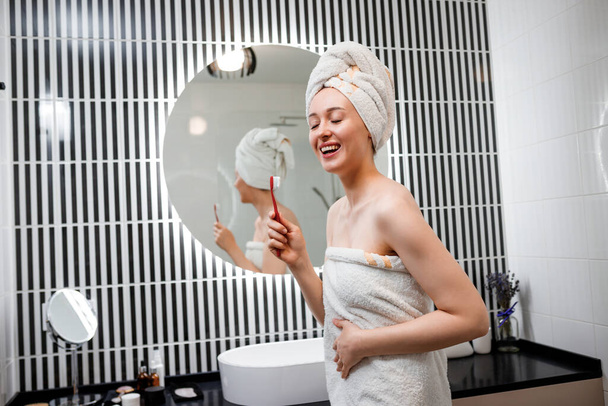Молодая счастливая женщина с закрытыми глазами в белом полотенце стоит в ванной комнате, держа зубную щетку и поет на нее, как в микрофоне - Фото, изображение