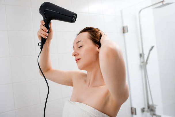 Young woman drying and styling hair with hair dryer, making hairstyle in modern bathroom interior. Beauty routine after morning shower - Фото, изображение