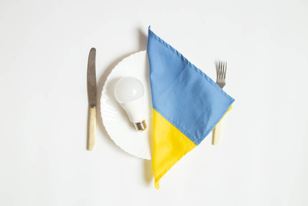 Светодиодная лампочка и флаг Украины лежат на обеденной тарелке и рядом с ножом с вилкой на белом фоне, отключения электроэнергии в Украине из-за войны - Фото, изображение