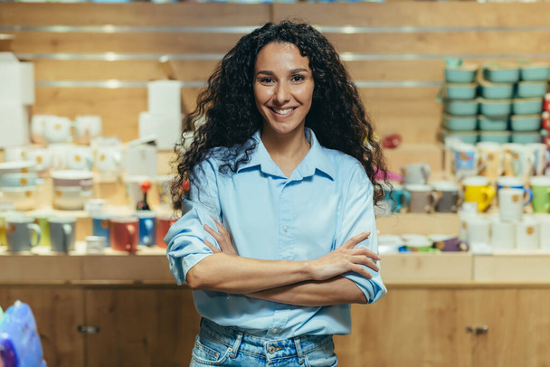 Portrait of business owner, hispanic woman managing gift shop, woman with curly hair looking at camera and smiling with arms crossed. - Foto, Imagen