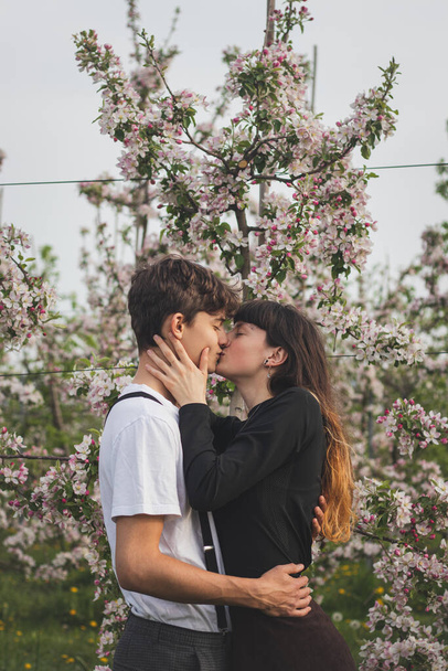 Νεαρή δυνατή αγάπη ανάμεσα σε δύο υπέροχους ανθρώπους που περπατούν κάτω από μηλιές. Απλό πορτραίτο ενός ζευγαριού με καθημερινά ρούχα. Φιλί αγνής αγάπης. - Φωτογραφία, εικόνα