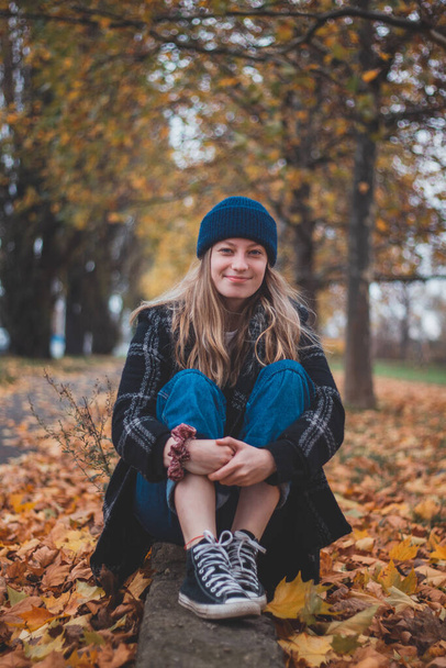 Smiling and happy brunette sitting in a pile of colourful leaves in a city park. Candid portrait of a young real woman in autumn clothes in the fall season. Orange, red and brown colour. - Photo, Image
